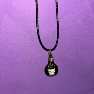 Svart halsband med katt i glödlampa - Unikt svart halsband med ett hänge format som en glödlampa. Inuti hänget finns en söt vit katt med stängda ögon och stjärnor runtomkring. Perfekt accessoar för dig som gillar katter och vill ha något annorlunda.