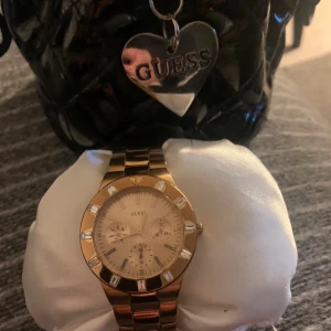 Guldig klocka från Guess - Stilren guldig klocka från Guess med länkarmband i metall och glittriga stenar runt urtavlan. 