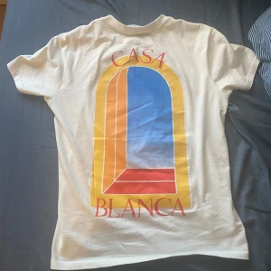 Creme färgad t-shirt från Casablanca - Snygg creme färgad t-shirt från Casablanca med färgglatt grafiskt tryck i gult, blått och rött på både bröstet och ryggen. Klassisk passform med rund hals och korta ärmar. Perfekt för dig som gillar stilrena men ändå unika plagg.