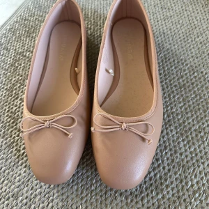 Beiga ballerinaskor med rosett - Snygga ballerinaskor i beige från Hush Puppies med rund tå och söt rosettdetalj framtill. Skorna har platt sula och är tillverkade i syntetmaterial, perfekta för en stilren och enkel look.