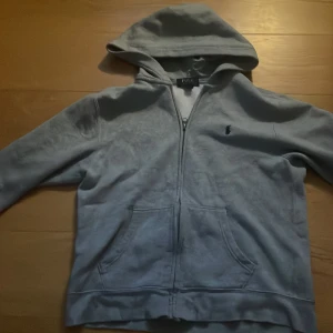 Grå hoodie från Polo Ralph Lauren - Grå hoodie från Polo Ralph Lauren med dragkedja och logga på bröstet. Bra använt skick! Säljer den för att den har växt ur mig( är lite liten för sin storlek)