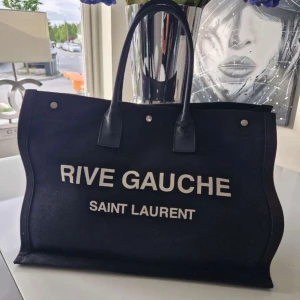 Rive Gauce Saint Laurent Totebag - Totebag med logotyp Tvả runda handtag, tryckknapp,logotyp tryck framtill, innerficka med dragkedja Bra skick- Sparsamt använd! Kvitto finns ! Inköpt fràn Fashionette!