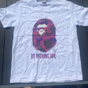 Vit BAPE t-shirt med lila camo-apa - Large - Vit t-shirt med ett stort lila kamouflagemönstrat A Bathing Ape-tryck på framsidan. Trycket föreställer en apa och texten ”BY BATHING APE”. Använd ca 5 gånger, tvättad sen dess. Storlek large (L)