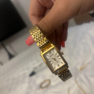 Guldig  klocka från Sontas - Stilren klocka från Sontas med rektangulärt ur i guld och silver. Armbandet är i metall med länkdesign och urtavlan är vit med svarta romerska siffror. Platsen är kvar på klockan