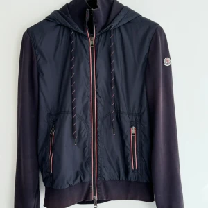 Äkta Moncler Cardigan - Blå vindjacka från Moncler med huva och dragkedja framtill. Jackan har två fickor med dragkedja och snygga röda detaljer längs dragkedjorna. Materialmix med glansig polyester framtill och mjukare tyg på ärmar och rygg. Klassisk Moncler-logga på ärmen. Säljer denna då den blivit för liten för mig. Storleken är L men den passar M. Cardiganen har små spår utav användning längst ner på armarna men som inte syns när man har på sig den. Säljer därför billigare. Äkta såklart.