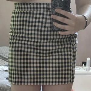 Rutig kortkjol i svart och beige - Supersnygg kortkjol med rutigt mönster i svart och beige. Kjolen har en tight passform och hög midja som ger en clean och trendig look. Perfekt att styla med en enkel topp eller oversized hoodie för en chill vibe.