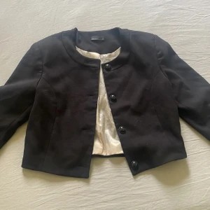 Svart croppad kavaj med knappar - Snygg svart croppad kavaj  med rundad krage och tre stora knappar framtill. Jackan har lång ärm och är fodrad med ljus insida. Perfekt för dig som vill ha en stilren och trendig look.