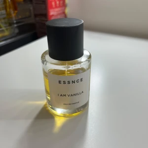 ESSNCE I Am Vanilla Eau de Parfum - Parfymflaska från ESSNCE med doften I Am Vanilla. Flaskan är cylindrisk i genomskinligt glas med svart matt kork och innehåller en gyllengul vätska. Minimalistisk vit etikett med svart text. Perfekt för dig som älskar söta och varma vaniljtoner.