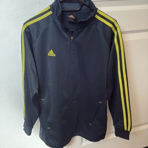 Adidas marinblå vindjacka med gula ränder - Snygg marinblå Adidas sweatshirt med klassiska gula ränder längs ärmarna och broderad logga på bröstet. Sweatshirten har dragkedja framtill, ribbade muddar och sportig look. Perfekt för dig som gillar retrostil och streetwear.