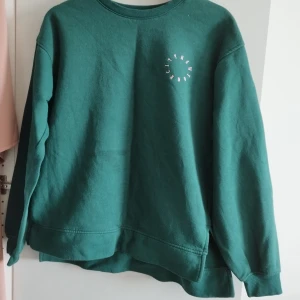 Grön sweatshirt New York City - Mörkgrön sweatshirt med rund hals och tryck i vitt och rosa på bröstet där det står 'New York City' i en cirkel. Tröjan har en loose passform och långa ärmar med ribbade muddar. Perfekt för en chill och avslappnad stil.