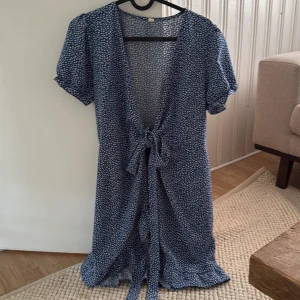 Blå långklänning med knyt och mönster - Långklänning i blått med vitt småmönster, korta puffärmar och djup v-ringning. Klänningen har knyt framtill i midjan och är tillverkad i ett lätt, flowy material som passar perfekt för varma dagar.
