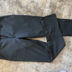 Svarta kostymbyxor från Grunt - Snygga svarta kostymbyxor från Grunt med klassisk rak passform. Använda 1 ggr på skolavslutningen. Byxorna har bälteshällor, dragkedja och knapp framtill. Perfekta för en clean och stilren look. Materialet känns mjukt och följsamt, vilket gör dem bekväma att bära hela dagen.