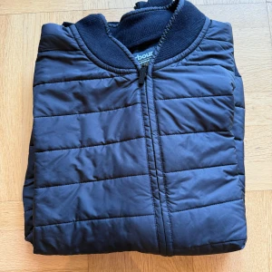 Svart quiltad cardigan/jacka Barbour - Snygg svart cardigan/jacka från Barbour International med quiltad framsida och stickade ärmar. Jackan har dragkedja framtill, ribbade muddar och Barbour-logga på ärmen. Perfekt för dig som gillar stilren och sportig look.