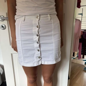 Vit jeanskjol med knappar från GinaTricot - Snygg vit kort jeanskjol från GinaTricot i storlek 36. Kjolen har raka linjer, bälteshällor och silverfärgade knappar framtill hela vägen ner. Perfekt för en fräsch och clean look i sommar.