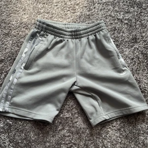 Grå Adidas shorts med ränder - Snygga grå shorts från Adidas med klassiska vita ränder längs sidorna och broderad logga framtill. Mjuk resår i midjan för skön passform. Perfekta för träning eller chill dagar. Sportig och stilren look som alltid funkar.