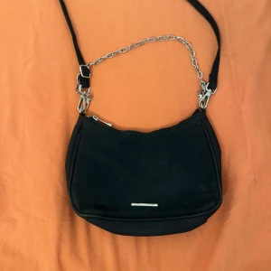 Svart axelväska från Bershka med kedja - Snygg svart axelväska från Bershka med silverfärgad kedjedetalj och justerbar rem. Väskan är tillverkad i syntetmaterial och har dragkedja upptill samt en liten innerficka. Perfekt storlek för det viktigaste och ger en cool vibe till din outfit.