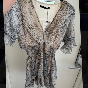 Zara kortklänning med leopardmönster - Supersöt kortklänning från Zara med ormmönster i beige, grått och svart. Klänningen har v-ringning, volangdetaljer och korta, fladdriga ärmar. Materialet är lätt och luftigt, perfekt för varma dagar. Smockad midja ger en snygg siluett. Aldrig används så är sågott som ny!