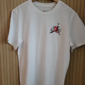Vit Nike Air Jordan t-shirt - Snygg vit t-shirt från Nike Air Jordan med ikoniskt Jumpman-tryck i svart, grått och rött på bröstet och stort tryck på ryggen. Klassisk passform och rund hals. Perfekt för dig som gillar streetwear och sportig stil.