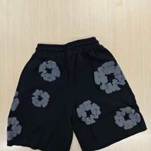 Svarta shorts från Denim Tears M - Coola svarta shorts från Denim Tears med unika gråa blommiga mönster över hela tyget. De har elastisk midja med snörning för skön passform. Perfekta för dig som vill sticka ut med en streetwear vibe och gillar statement-plagg.