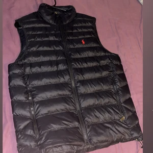 Svart dunväst från Polo Ralph Lauren - Snygg svart dunväst från Polo Ralph Lauren med klassisk röd logga på bröstet. Västen har quiltad design, hög krage och dragkedja framtill samt två praktiska fickor med dragkedja. Perfekt för lager-på-lager och streetstyle.