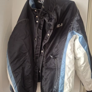 Svart och vit pufferjacka från FILA - Säljer en fet pufferjacka från FILA i svart med vita och ljusblå detaljer. Jackan har dragkedja, tryckknappar och stora loggor både fram och bak. Perfekt för dig som vill sticka ut med en sportig vibe och hålla dig varm under kalla dagar.