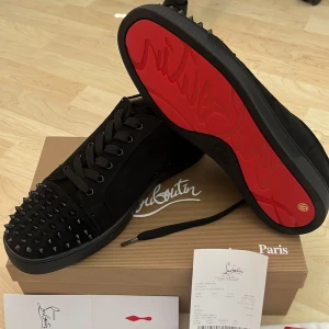 Christian Louboutin svarta sneakers med nitar - Snygga svarta sneakers från Christian Louboutin med ikonisk röd sula och svarta nitar på tån. Skorna har snörning och är tillverkade i mocka och skinn. Perfekt för dig som vill sticka ut med en lyxig och edgy stil.
