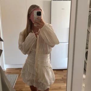 Vit omlottklänning med volanger - Superfin kort vit klänning med omlottknytning i halsen, långa puffiga ärmar och volangkjol. Klänningen har markerad midja och är gjord i ett lätt, glansigt material som ger en lyxig känsla. Perfekt för sommarens alla tillfällen! Lappen finns kvar och den är helt ny.