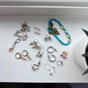 Smyckesmix smått & gott - Flera örhängen och halsband i silver- och guldfärg, vissa med pärlor och stenar. Ett armband med turkosa pärlor och guldiga blommor. Mix av små studs, hoops och ett halsband med rosa sten. Perfekt för dig som gillar att variera din stil. Skickas i en Lily & Rose-ask.