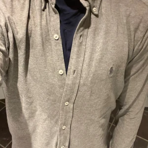 Grå skjorta från Ralph Lauren - Säljer en stilren grå skjorta från Ralph Lauren i featherweight mesh. Skjortan har långa ärmar och knappar framtill. Den klassiska loggan är broderad på bröstet. Perfekt för en avslappnad men ändå elegant look.