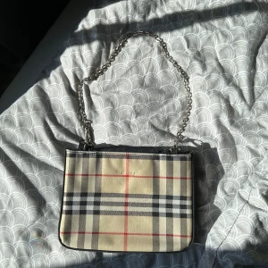 Rutig axelväska med kedja - Snygg axelväska i klassiskt beige, svart och rött rutigt mönster. Väskan har en silverfärgad kedjerem och dragkedja både på insidan och utsidan. Perfekt storlek för det viktigaste och har flera fack för bra organisering. Tyvärr ett litet hål på insidan, men inget som stör! 