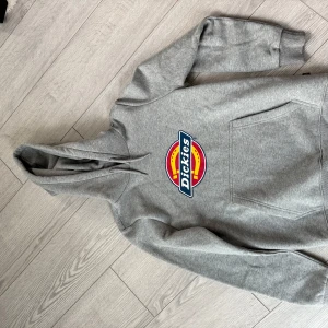 Grå Dickies hoodie med logga - Säljer en grå hoodie från Dickies i storlek small. Tröjan har stor färgglad Dickies-logga på bröstet, huva med dragsko och en klassisk magficka. Tillverkad i mjuk bomullsblandning, perfekt för chill dagar eller streetwear-stil.