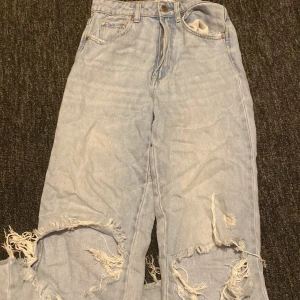 Ljusa slitna jeans med hål - Säljer ett par ljusblå jeans med stora slitningar och hål vid knäna. Jeansen har klassisk femficksdesign, dragkedja och knapp framtill. Perfekta för en avslappnad och trendig streetstil.
