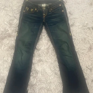 True Religion bootcut jeans blå, stl 26 - Snygga blå jeans från True Religion med bootcut passform och coola kontrastsömmar. Bakfickorna har den klassiska gula brodyren och loggan. Jeansen har en låg midja och är tillverkade i denimtyg. Knapparna är lite rostiga men inget man märker från långt hål finns små skador på jeansen men ingen man lägger märke till!