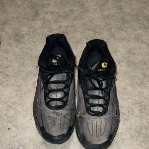 Nike Air Max Plus Tn i svart och grå - Nike Air Max Plus Tn sneakers i svart och grå med klassisk Tn-logga på plösen. Skorna har svarta snören, mesh- och syntetpaneler samt Tuned Air-dämpning i sulan. Snygga detaljer med reflekterande inslag och en sportig vibe. Perfekta för dig som gillar streetwear.