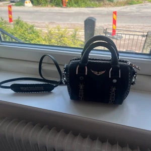 Svart Zadig & Voltaire väska - Cool svart handväska från Zadig & Voltaire i skinn med silverfärgade nitar, kedjedetaljer och logga framtill. Väskan har både handtag och axelrem med broderad VOLTAIRE-text. Säljer för att jag redan har så många väskor!💕💕💕Väskan är i fint skick men som man ser på sista bilden har själva dragkedjan rostat lite men det är inte man tänker på!🥰🥰Säljer för bra pris💕💕