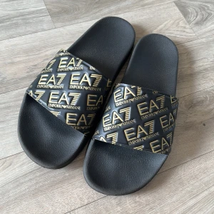 Svarta slides från Emporio Armani EA7 - Svarta slides från Emporio Armani EA7 med bred rem i svart och guld där loggan EA7 Emporio Armani upprepas över hela remmen. Snygg och sportig design, perfekt för sommaren eller att chilla i hemma.