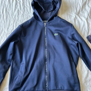 Mörkblå hoodiejacka från Lacoste Sport - Mörkblå hoodiejacka från Lacoste Sport med dragkedja och broderad krokodil på bröstet. Jackan har huva, två fickor och är helfärgad. Insidan är mjuk och fleece-liknande, perfekt för kyliga dagar. Snygg och sportig stil med Lacoste-logga längs dragkedjan.