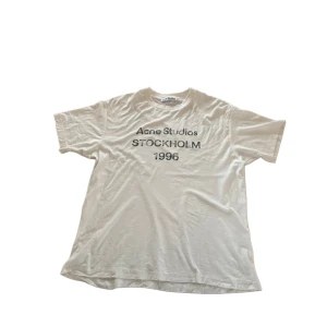 Vit t-shirt Acne Studios Stockholm - Vit t-shirt från Acne Studios med svart tryck på bröstet där det står 'Acne Studios Stockholm 1996'. Klassisk rund halsringning och korta ärmar. Skön och enkel modell i mjuk bomull, perfekt för en clean och avslappnad look.