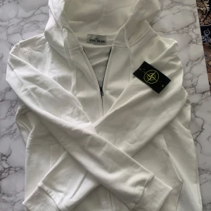 Vit hoodie från Stone Island - Snygg vit hoodie från Stone Island med dragkedja och klassisk logga på ärmen. Tröjan har huva, långa ärmar och är tillverkad i mjuk bomull. Perfekt för dig som gillar streetwear och vill ha en clean look. Inga repor eller skadade.
