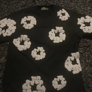Svart t-shirt Denim Tears XL - Svart t-shirt från Denim Tears i storlek XL med unika vita blommiga tryck över hela tröjan. Klassisk passform och korta ärmar. Tillverkad i USA, perfekt för dig som gillar streetwear med statement-mönster.