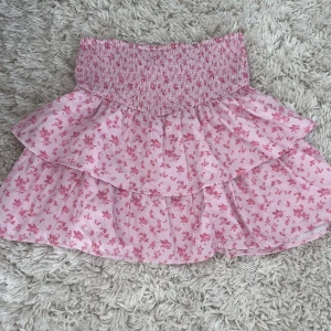 Rosa blommig volangkjol Gina Tricot - Supersöt rosa kortkjol från Gina Tricot med blommigt mönster och bred smock i midjan. Kjolen har dubbla volanger som ger en fluffig och lekfull look. Perfekt för dig som gillar pastell och romantiska vibes. Storlek 146/152 passar XS