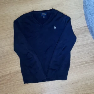 Mörkblå stickad tröja Polo Ralph Lauren - Mörkblå stickad tröja från Polo Ralph Lauren med klassisk v-ringning och broderad logga på bröstet. Tröjan har långa ärmar och ribbade muddar vid ärmslut och nederkant. Perfekt för dig som gillar stilren och tidlös design.