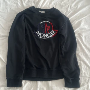 Svart Moncler sweatshirt med stor logga - Svart sweatshirt från Moncler med stor broderad logga i vitt, rött och blått på bröstet. Tröjan har rund hals, ribbade muddar och är långärmad. Tillverkad i mjuk bomull som känns skön mot huden. Perfekt för dig som gillar streetwear med lyxig vibe.