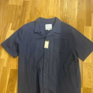 Mörkblå kortärmad skjorta med struktur - Snygg mörkblå skjorta från lager 152 med korta ärmar och diskret strukturerat mönster. Skjortan har klassisk krage, bröstficka och knäppning framtill. Perfekt för en stilren och avslappnad look. Säljs då det inte var min still