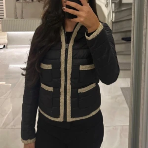 Svart quiltad pufferjacka med beige detaljer - Säljer en svart quiltad pufferjacka med beige kantdetaljer längs dragkedja, ärmslut, fickor och nederkant. Jackan är croppad och har lång ärm, dragkedja framtill och fyra fickor framtill. Perfekt för dig som vill ha en stilren men unik look.
