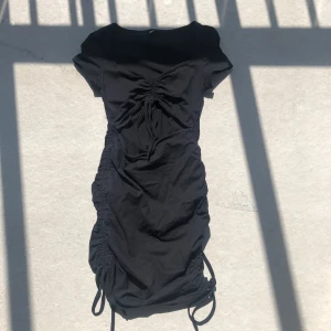 Svart kort klänning med snörning - Svart kortklänning i bomull med snygg snörning framtill och rynkade detaljer längs sidorna. Klänningen har korta ärmar och en v-ringad halsringning. Perfekt för dig som gillar en tight och trendig look.