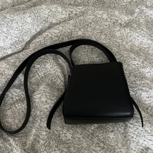 Svart axelväska  - Säljer en stilren svart axelväska med enkel och modern form. Väskan har långa axelband och är tillverkad i ett slätt material som ger en clean look. Perfekt för dig som gillar minimalistiska accessoarer och vill ha något som funkar till många outfits. Ifrån ginatricot