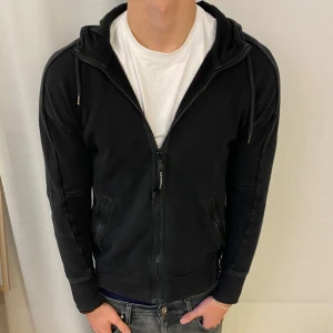 Svart hoodiejacka från C.P. Company - Svart hoodiejacka från C.P. Company med dragkedja framtill, två fickor med dragkedja och ikoniska goggles i huvan. Den är i topp skick och bara använd ett få antal gånger , skriv för frågor eller likande 🤗
