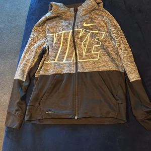Nike Dri-Fit hoodie i svart och grå - Nike Dri-Fit hoodie med dragkedja, stor logga framtill och huva. Jackan är svart nertill och gråmelerad upptill, med långa ärmar och två fickor. Perfekt för träning eller chill, tillverkad i funktionsmaterial som andas.