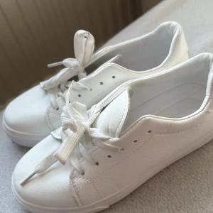 Vita sneakers i skinnimitation - Fräscha helvita sneakers med klassisk design och rund tå. Skorna har breda vita snören och är gjorda i skinnimitation med diskreta sömmar. Perfekta för en clean och stilren look.